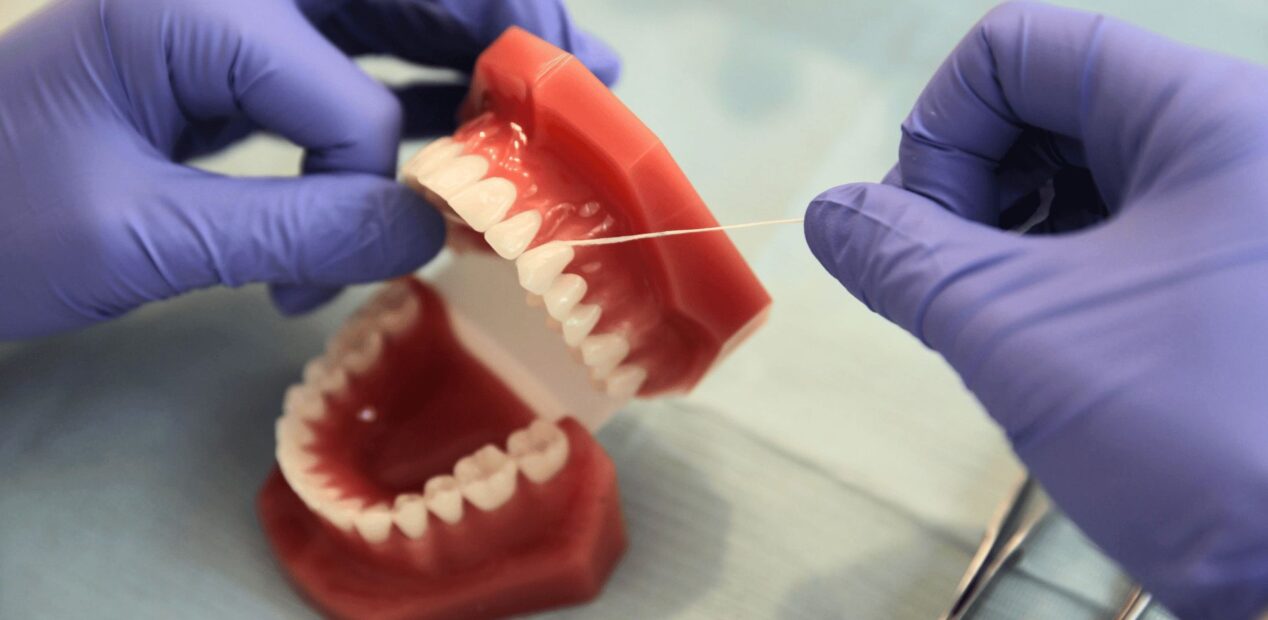Une personne portant des gants violets au iDental Studio démontre l'utilisation de fil dentaire sur un modèle en plastique de dents humaines, avec des outils dentaires visibles à proximité sur une surface bleue dans le cabinet dentaire.