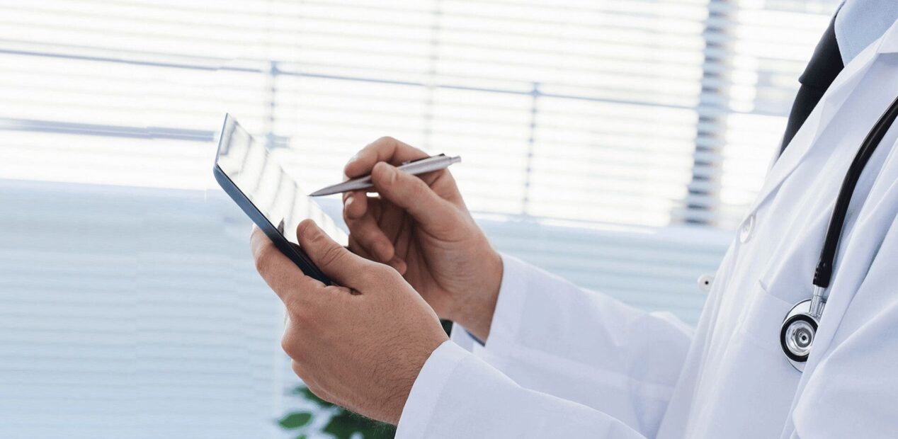 Un médecin en blouse blanche et stéthoscope tient une tablette numérique, utilisant un stylet pour écrire ou naviguer - une image reflétant l'atmosphère moderne d'un cabinet dentaire iDental Studio, avec des stores et une lumière vive en arrière-plan.