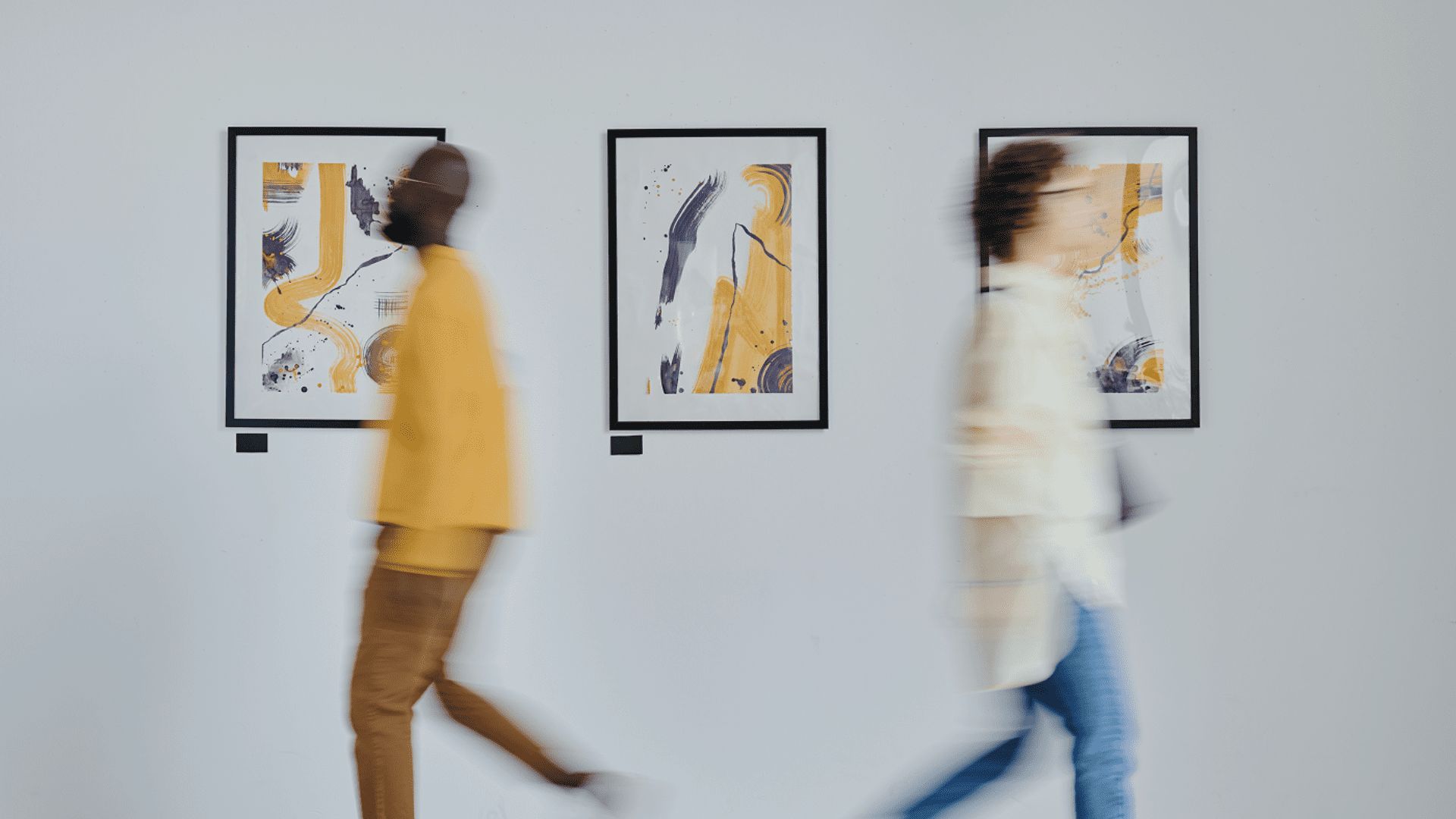 Two people walk past abstract art paintings framed on a white gallery wall at iDental Studio dental practice in Geneva. The figures are blurred in motion, while the artwork features bold yellow, black, and white shapes and lines.