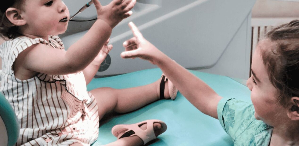 Two young children are smiling and reaching toward each other while one sits on a teal dental chair, holding a dental instrument, in the brightly lit iDental Studio dental practice in Geneva.