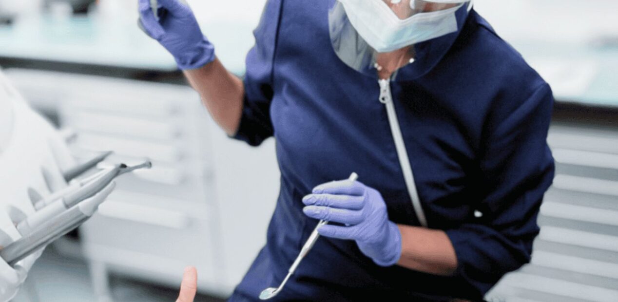 Au iDental Studio, un dentiste portant des gants, un masque et des lunettes de protection soigne un patient. Ce dernier, détendu dans son cabinet dentaire, muni d'un ouvre-bouche et de lunettes de sécurité orange, lève le pouce en signe d'approbation.