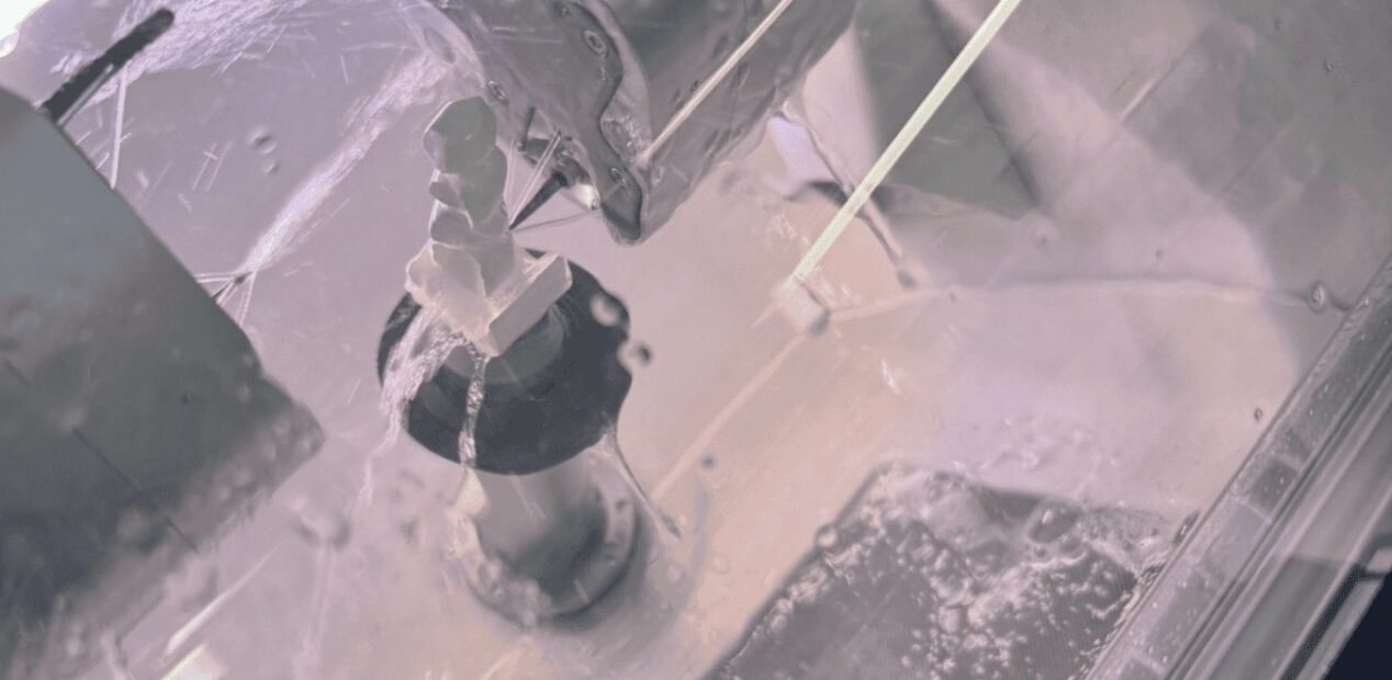 Vue rapprochée d'un dispositif mécanique pulvérisant de l'eau dans un environnement contrôlé chez iDental Studio, avec des gouttelettes d'eau et des éclaboussures visibles sur la surface transparente et les composants métalliques.