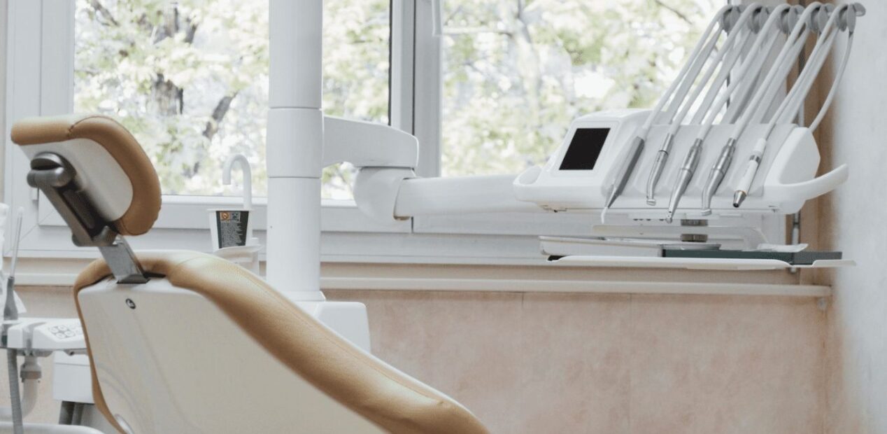 Un fauteuil dentaire moderne au revêtement beige est installé près d'une fenêtre du cabinet dentaire iDental. On peut y voir des instruments dentaires et une lampe d'examen suspendue, tandis que des arbres feuillus sont visibles à l'extérieur de la fenêtre lumineuse du cabinet dentaire.