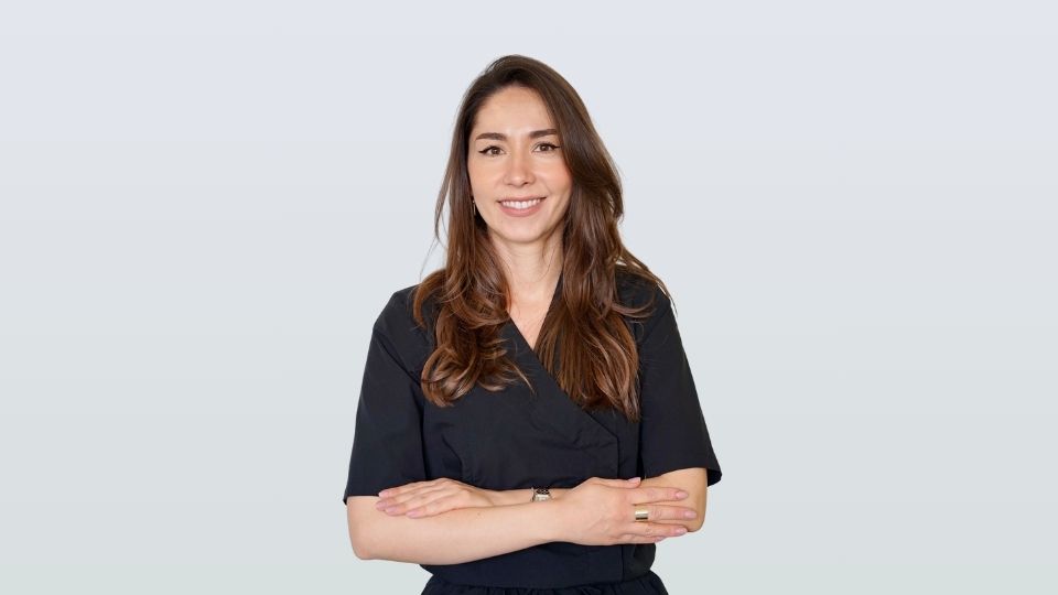 Dr Ioana Puicescu portant un haut noir à manches courtes se tient debout, les bras croisés, souriant à la caméra sur un fond clair et uni dans l'atmosphère accueillante d'iDental Studio.