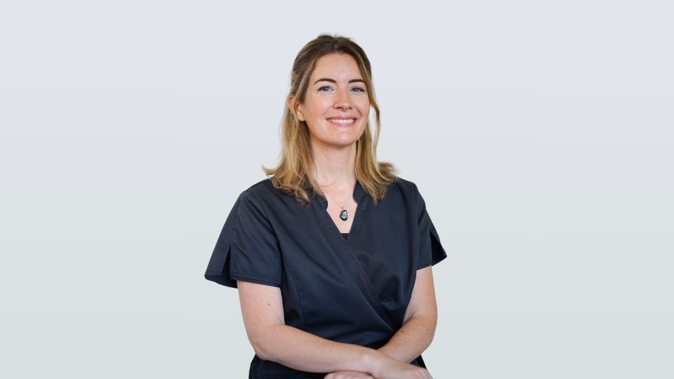 Laurie, portant une blouse médicale foncée et un collier, est assise en souriant sur un fond uni et clair, incarnant l'atmosphère accueillante du cabinet dentaire iDental Studio.