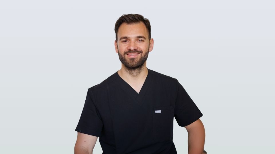 Dr Octavian Isac, vêtu d'une blouse médicale noire, sourit assis devant un fond gris clair uni au cabinet dentaire iDental Studio.